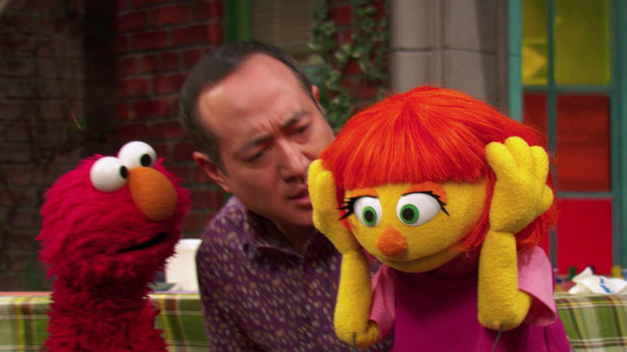 Julia covers her ears as loud siren sounds, and Alan explains to the other Muppets that she doesn’t like loud noises. Image credit: Goodstuffbuzz.com